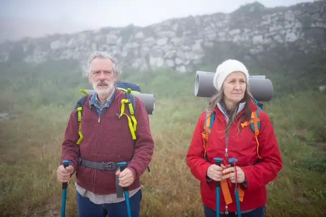 A gray-haired couple in hiking gear with backpacks and trekking poles on a misty trail