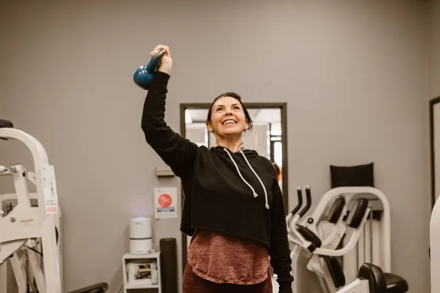 A smiling middle-aged woman in a black hoodie lifting a kettlebell overhead in a small gym